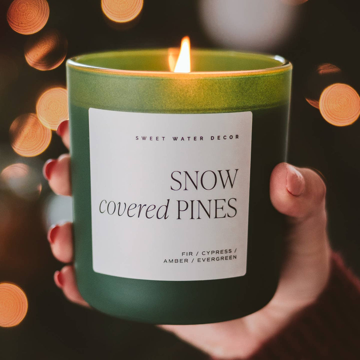 Hand holding a 'Snow Covered Pines' candle with a blurred light background