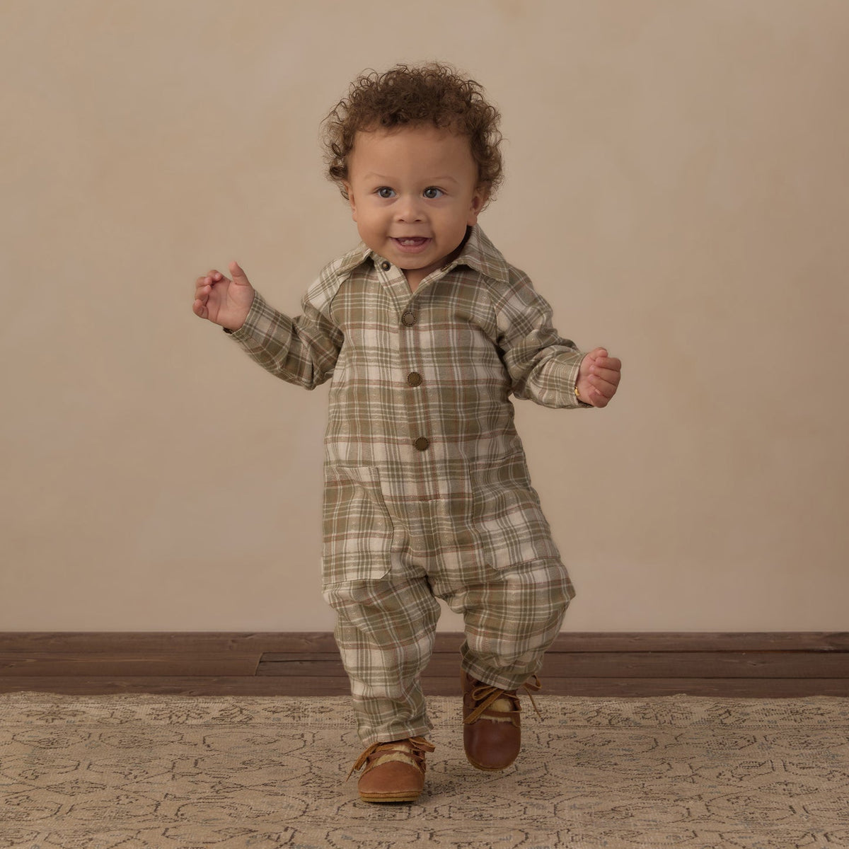 A toddler with curly brown hair wearing a beige and brown plaid button-up romper and brown leather shoes, walking forward on a patterned rug against a beige studio backdrop.