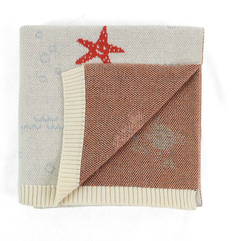 A folded knit blanket with a warm brown center, cream ribbed borders, a red starfish motif, and faint blue nautical patterns on a white background.