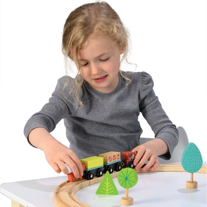 A young child playing with the My First Wooden Train Set by Mentari, assembling the wooden tracks and setting up a toy train, house, and trees on a white surface.