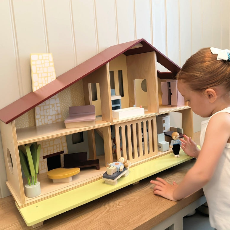 A young child wearing a white dress with a hair bow plays with the Wooden Cedar Chalet Modernist Dollhouse by Mentari, leaning over the open-front mid-century modern home while arranging furniture and dolls inside and on the attached patio.