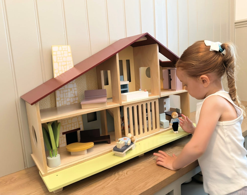 A young child wearing a white dress with a hair bow plays with the Wooden Cedar Chalet Modernist Dollhouse by Mentari, leaning over the open-front mid-century modern home while arranging furniture and dolls inside and on the attached patio.