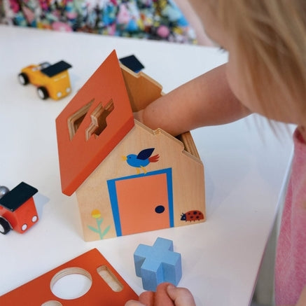 A toddler sits on the floor, holding a blue wooden puzzle piece while exploring a wooden shape sorter house and various colorful geometric blocks scattered nearby.