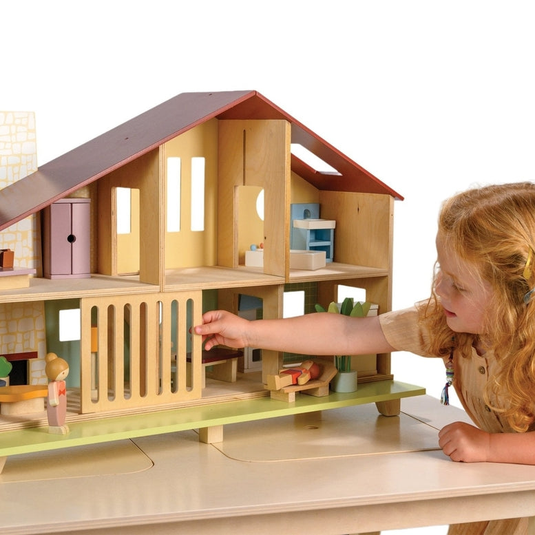 A young child with curly hair and a yellow dress plays with the Wooden Cedar Chalet Modernist Dollhouse by Mentari, sliding open the ground-floor grill on the mid-century modern home while arranging miniature furniture and dolls inside and on the patio.