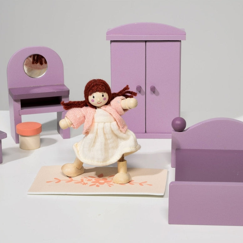 A dollhouse bedroom scene featuring a wooden doll in a pale dress holding a small bag, standing near the center in front of a purple double bed. In the background, a matching purple wardrobe stands beside a dressing table with an oval mirror and a matching stool, while a bedside table with an orange base and yellow shade sits to the left.
