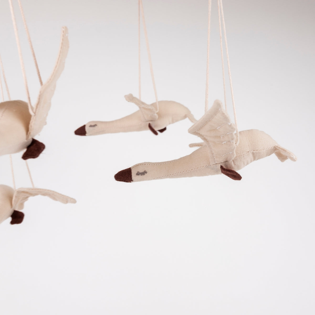 Three cream-colored fabric swan ornaments with dark brown beaks and feet, suspended by thin strings from a mobile against a plain white background.
