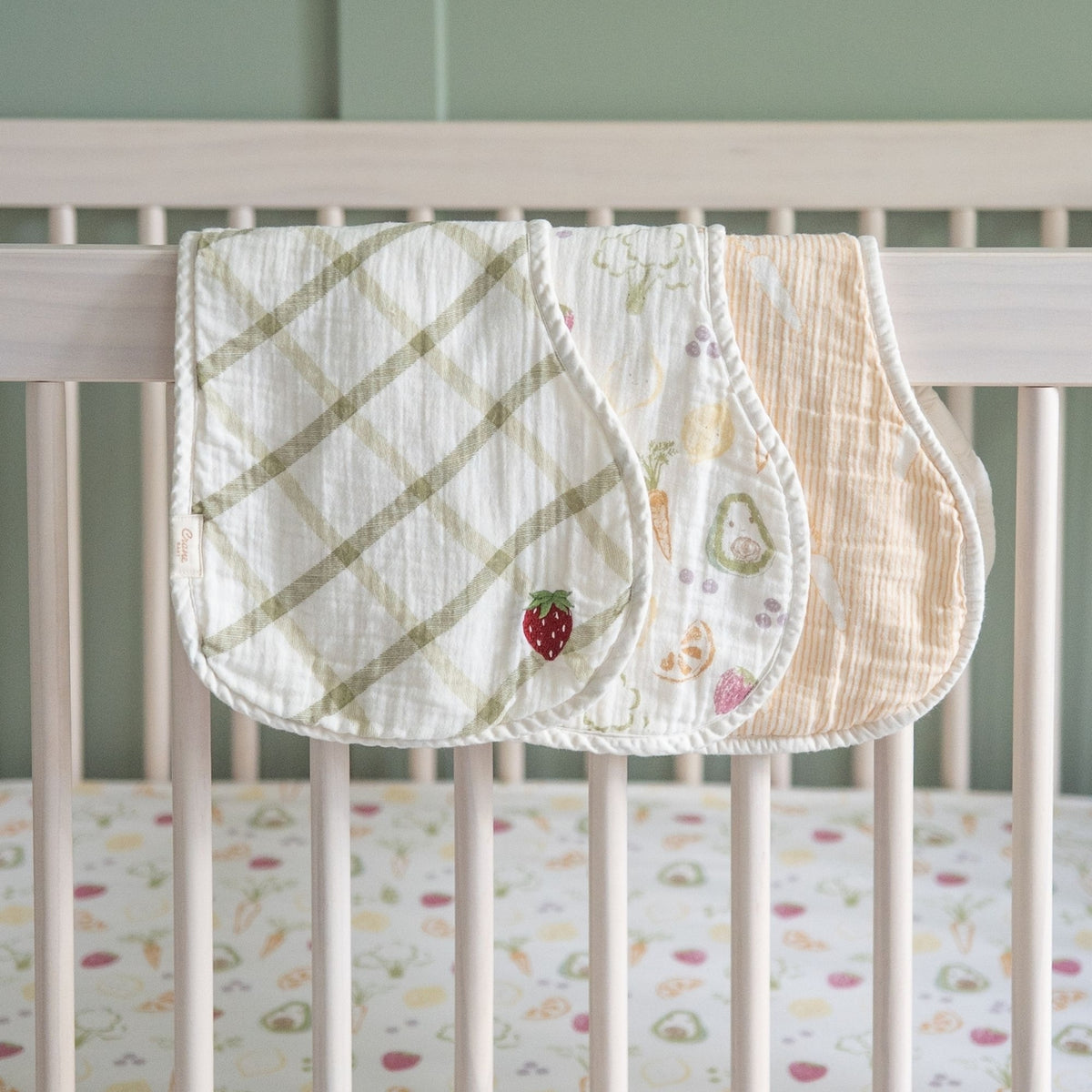 Three soft muslin baby blankets in different patterns and colors are draped over the side of a light wood crib. The left blanket has a sage green plaid pattern, the middle has a white background with a small strawberry and green leaf motif, and the right blanket is a warm beige with a subtle floral print.