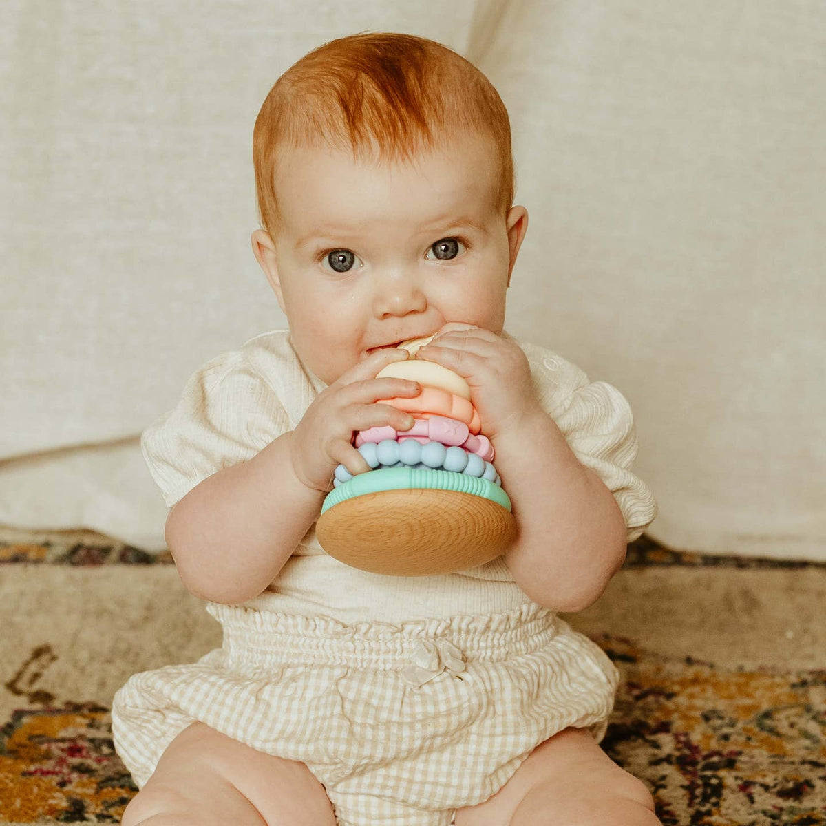 A baby with red hair wearing a cream-colored outfit is sitting on a patterned rug, holding and biting a wooden stacking toy with pastel-colored rings.