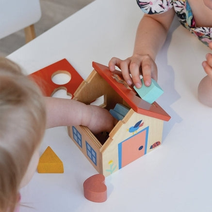 A young child looks down intently as they play with a wooden shape sorter house on a table, with an adult or older child leaning in beside them to watch. Colorful wooden blocks are scattered on the surface near the toy.