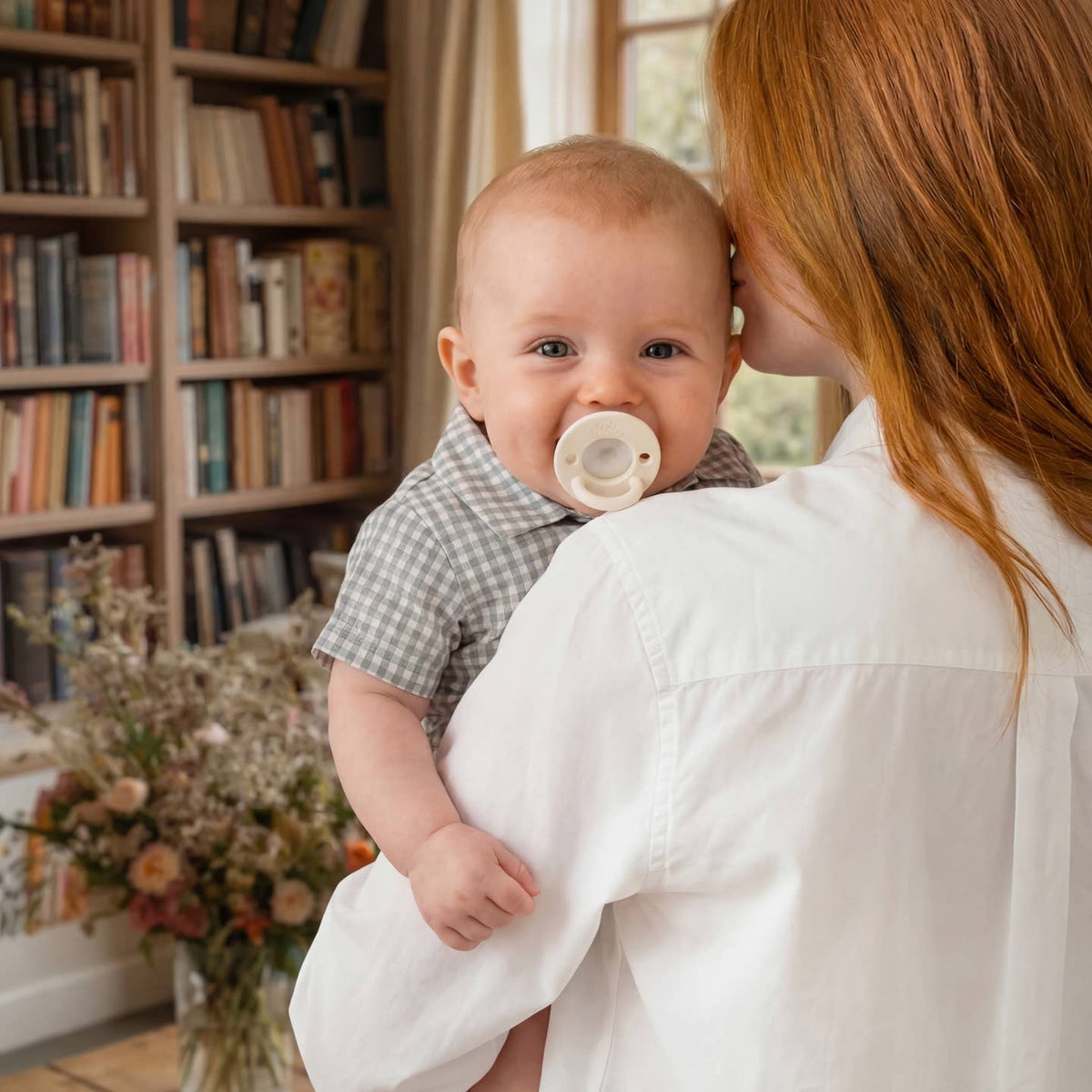 Woman holding a baby with a pacifier in a room with bookshelves and flowers.