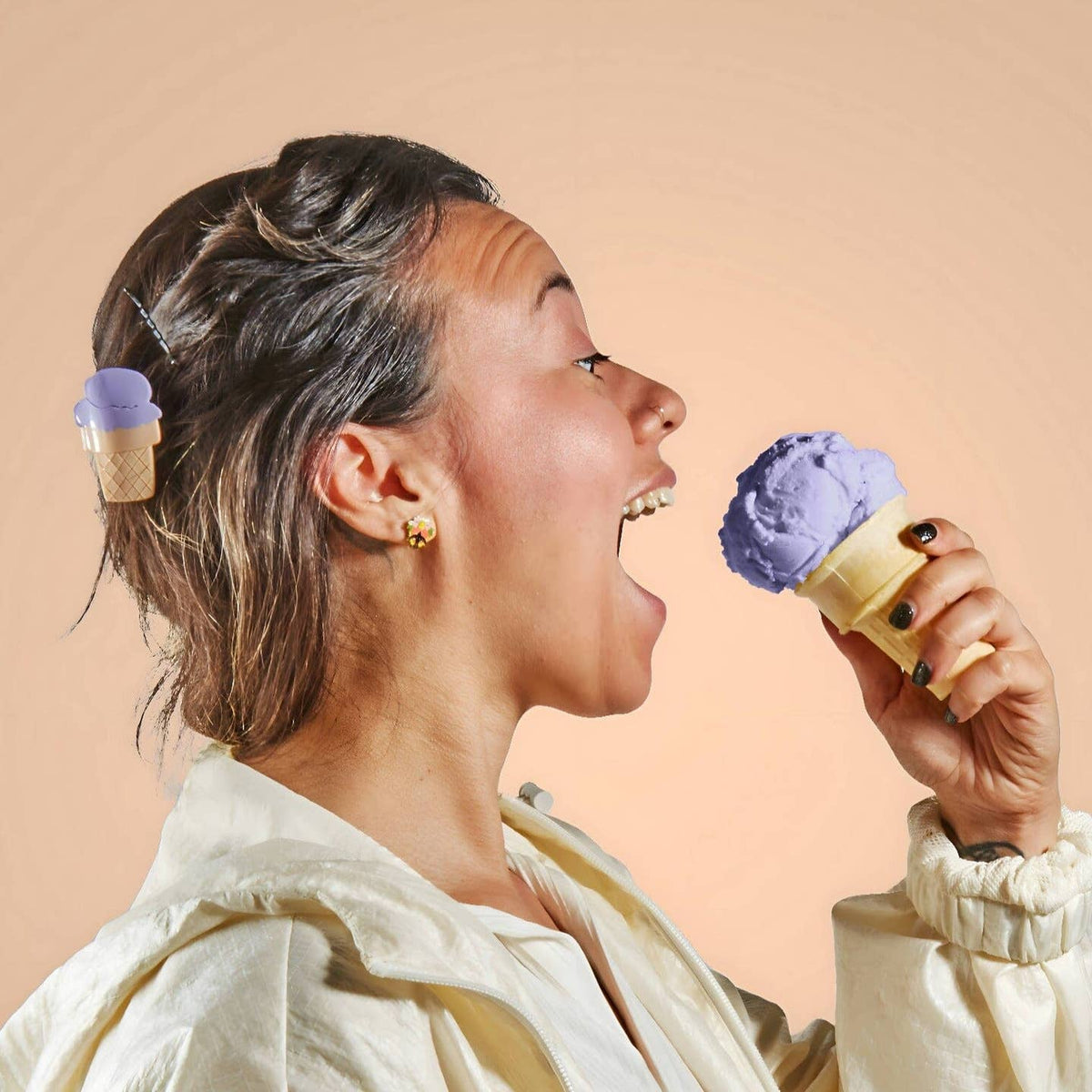 Woman eating a purple ice cream cone with another purple ice cream cone in her hair against a beige background