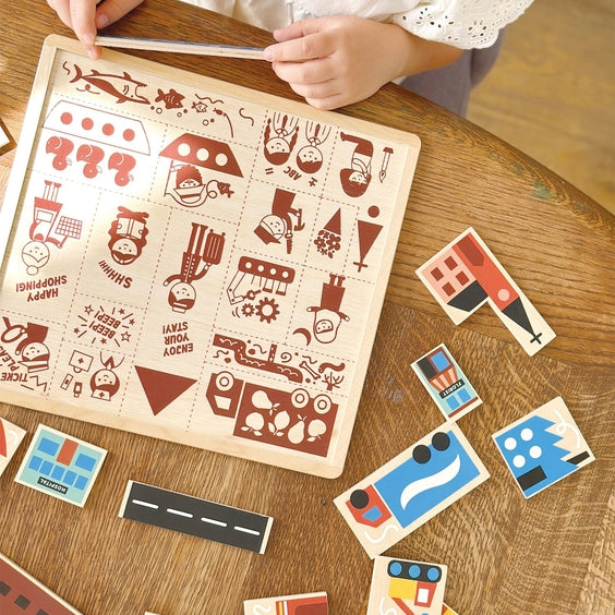 A child's hands hover above the Mentari Wooden Town Puzzle, arranging the pieces onto the illustrated wooden board on a tabletop.