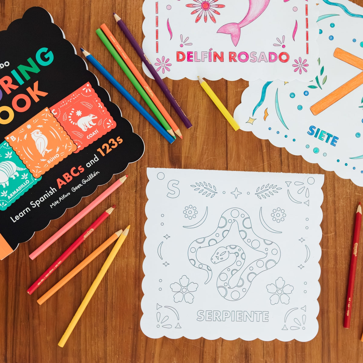 Spanish alphabet coloring book set with colored pencils and sample coloring cards arranged on a wooden surface. The set includes a black cover book labeled 'Nube Picasso' and white coloring pages with decorative Mexican-style borders.