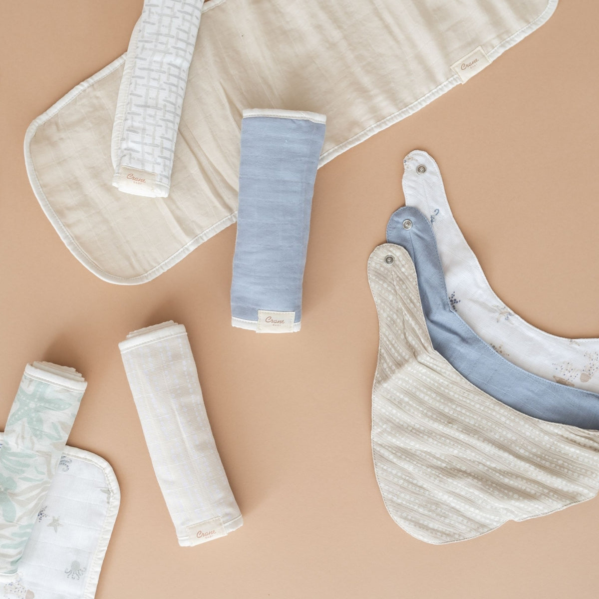 A styled flat-lay of baby muslin textiles on a warm beige background, including folded and rolled bibs and swabs in cream, light blue, and pale mint tones.