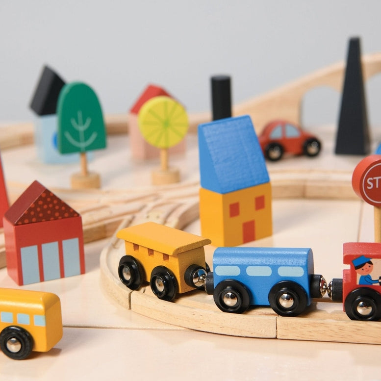 A close-up, angled view of a wooden toy train set featuring a red engine with a driver, a blue passenger car, and a yellow cargo car resting on wooden tracks. A round red "STOP" sign stands beside the track, and the background is filled with colorful wooden town elements, including a red house with a window, a yellow building, a blue bridge, and abstract green trees.