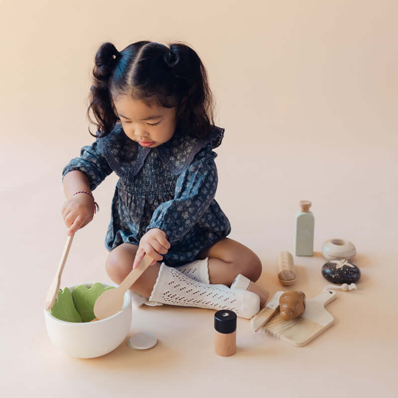 A young child with dark hair in pigtails sits on a beige floor, playing with a wooden toy kitchen set. The set includes a white bowl with green leaves, wooden spoons, small wooden bottles, a cutting board, and other play accessories.