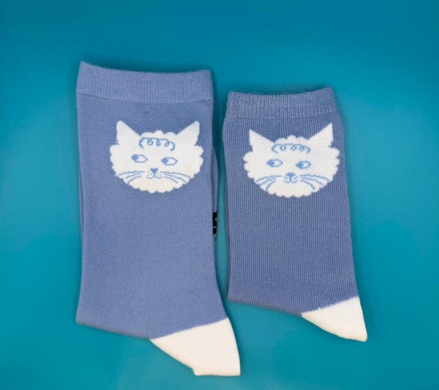 A pair of light blue ribbed socks with white cat faces and white toes, displayed on a vibrant teal background.