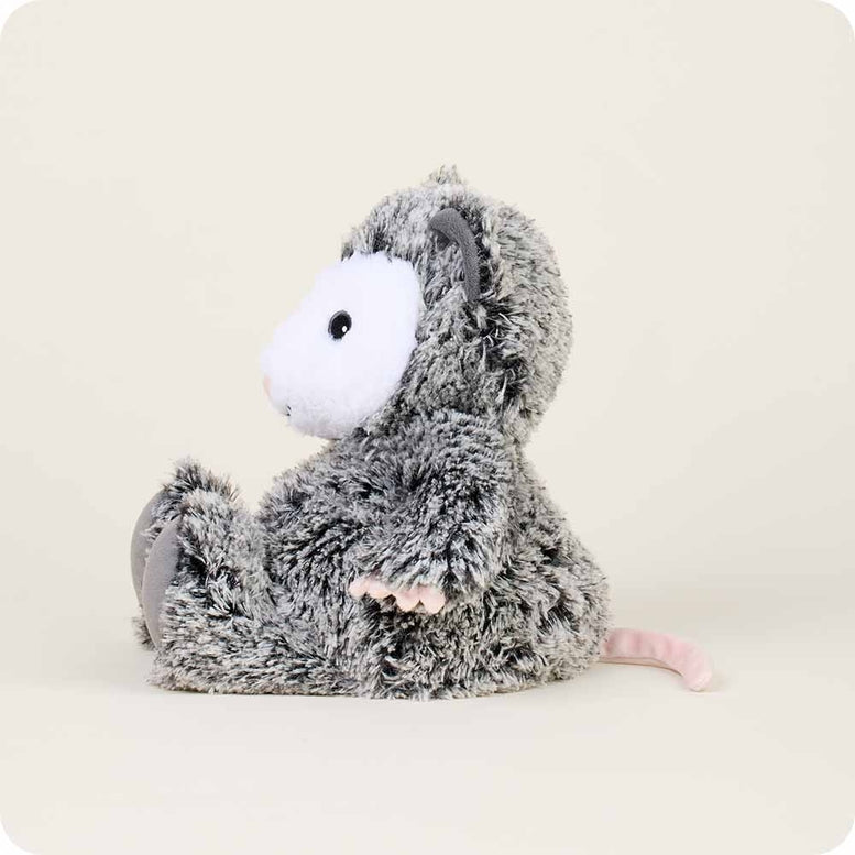 A gray and white fluffy Opossum by Warmies stuffed animal shown in profile, sitting sideways with its pink nose, white face, dark gray paws, and long pink tail visible against a neutral background.