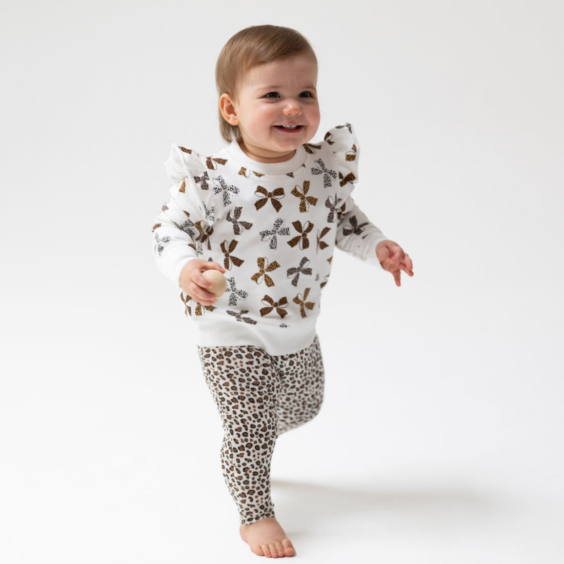 A smiling toddler walking barefoot on a white studio background, wearing a white long-sleeve top with brown bow prints and leopard-print leggings.