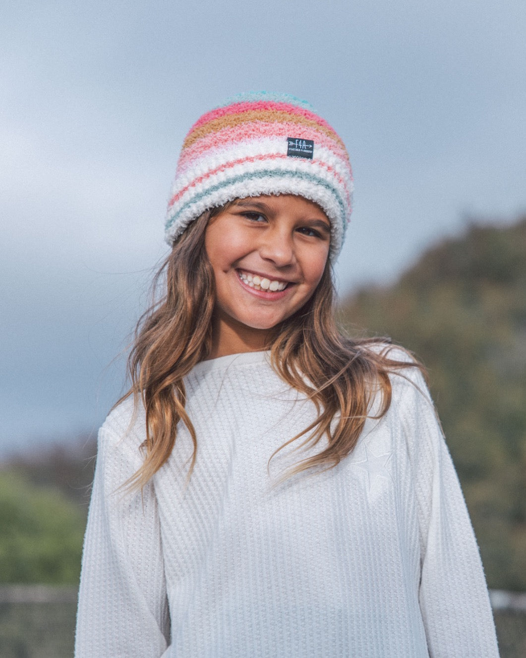 A smiling young girl wearing a white waffle-knit sweater and a colorful striped knit beanie hat with a small black brand patch, photographed outdoors against a soft-focus sky and trees.