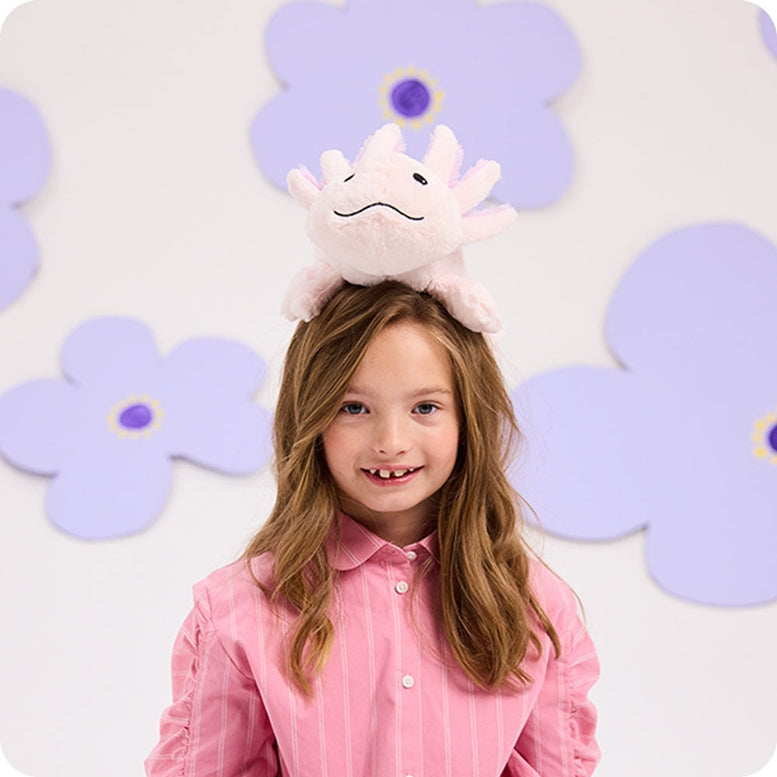 A young girl with long brown hair wearing a pink button-down shirt, balancing the Pink Axolotl plush toy on her head against a purple and white flower-patterned backdrop.