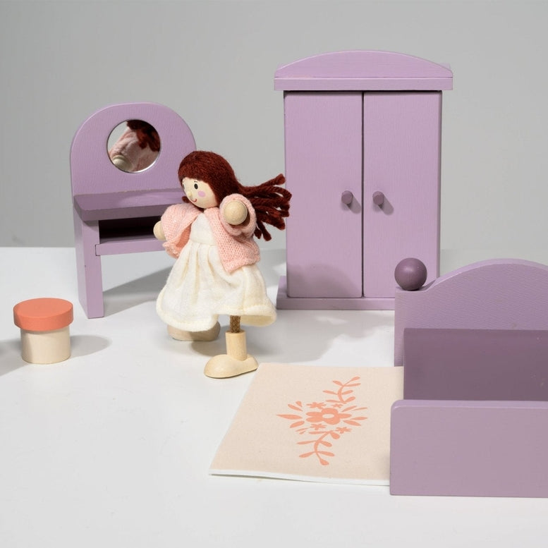 A dollhouse bedroom scene featuring a wooden doll in a pale dress standing near the center, positioned in front of a purple double bed. In the background, a matching purple wardrobe stands beside a dressing table with an oval mirror and a stool, while a bedside table with an orange-based lamp topped with a yellow shade sits on the left.
