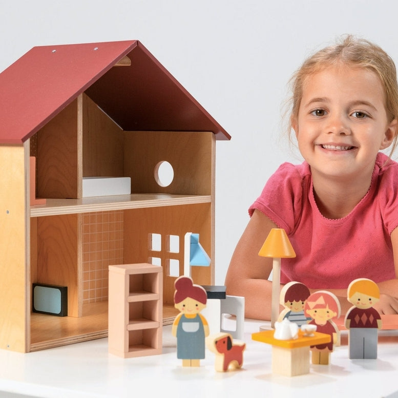A young child playing with an open-faced wooden dollhouse by Mentari, arranged on a white table with miniature wooden furniture and small figurines.