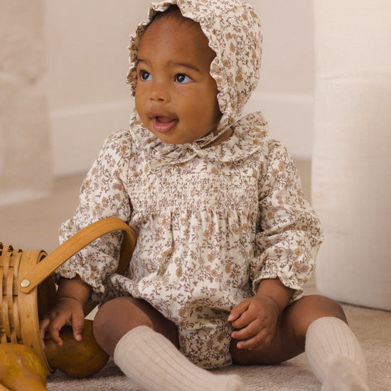 A baby wearing a cream and brown floral smocked romper with a matching ruffled bonnet, sitting on a carpeted floor next to a wicker basket.
