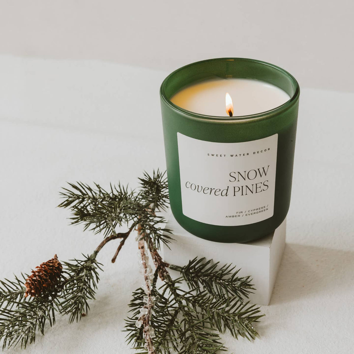 Candle labeled 'Snow Covered Pines' with a green lid and label, surrounded by pine branches on a light background.