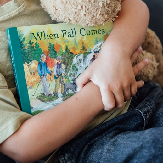 A toddler holding an open board book titled When Fall Comes, looking down at the colorful illustrated pages showing autumn trees and an outdoor nature scene.