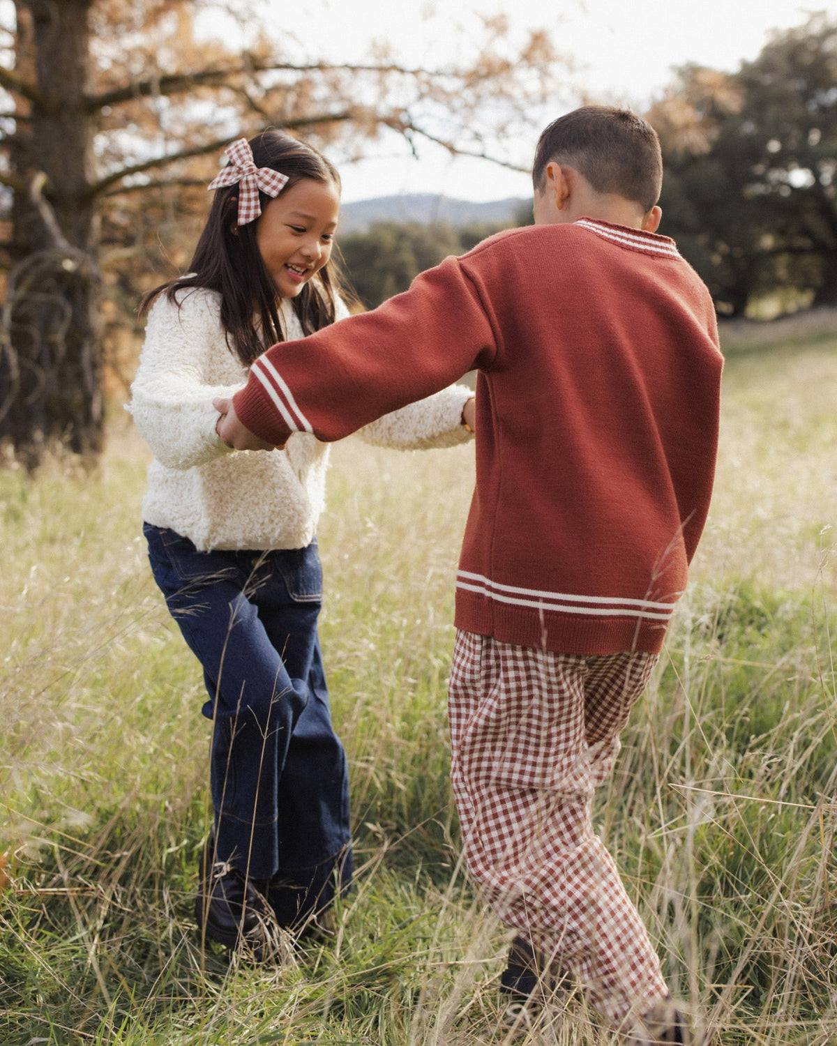 Two children playing in a grassy outdoor field. The child on the left wears a white textured sweater, dark blue jeans, and a red-and-white gingham bow in her hair.