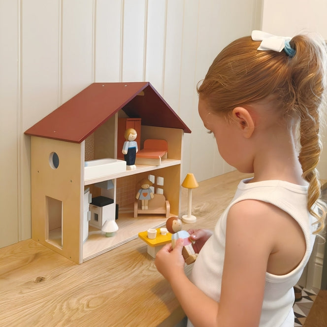 A young child playing with an open-faced wooden dollhouse by Mentari, arranged on a white table with miniature wooden furniture and small figurines.