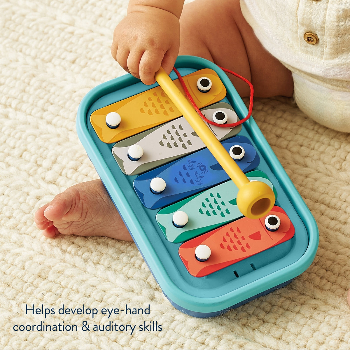 Colorful toy xylophone with a child's hand holding a mallet on a light-colored carpet.