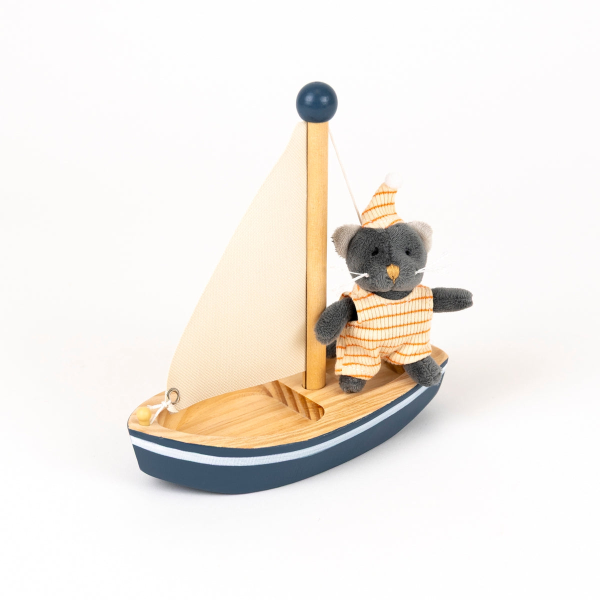 A wooden toy sailboat with a small gray plush cat character standing on the deck, wearing an orange-and-white striped outfit and matching hat, photographed on a clean white background.