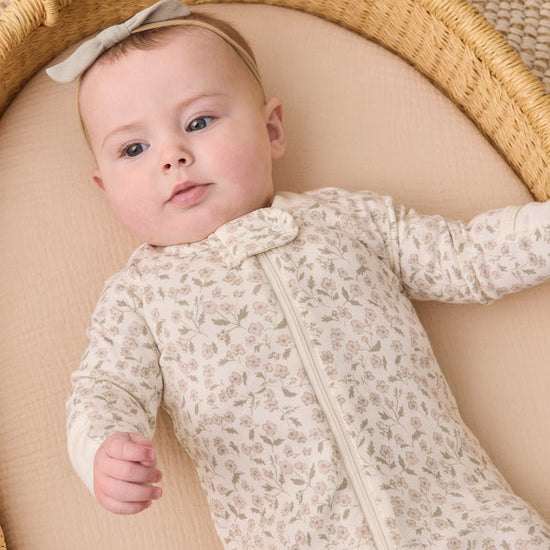 A baby lying in a woven bassinet wearing a cream-colored long-sleeve footless sleeper with a delicate floral pattern and a matching bow headband.