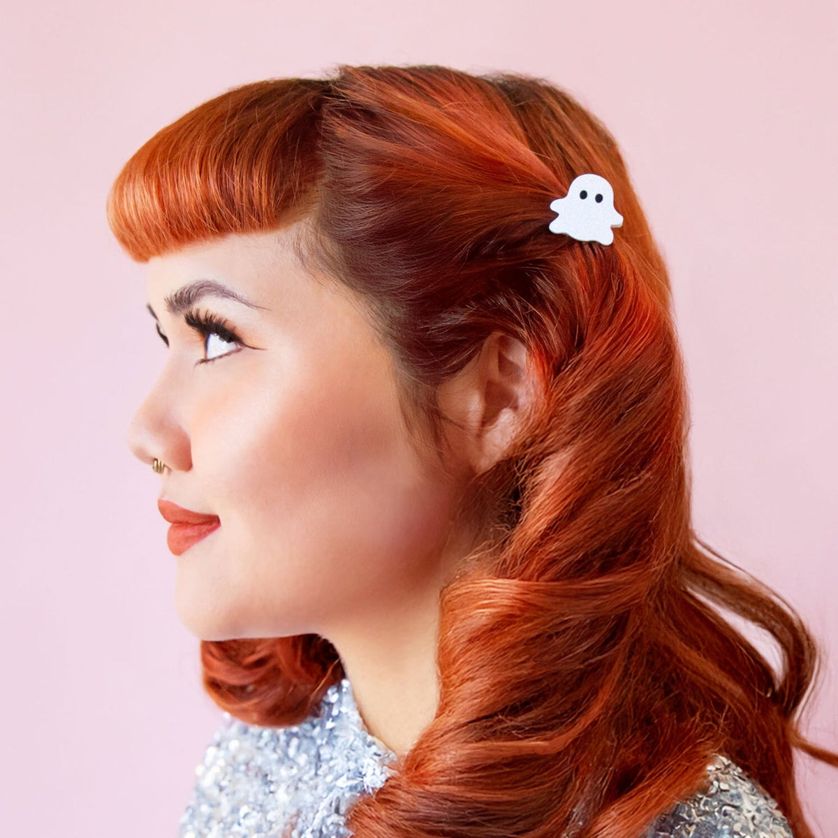 A woman with styled red hair shown in profile, wearing a small white ghost-shaped hair clip against a soft pink background.