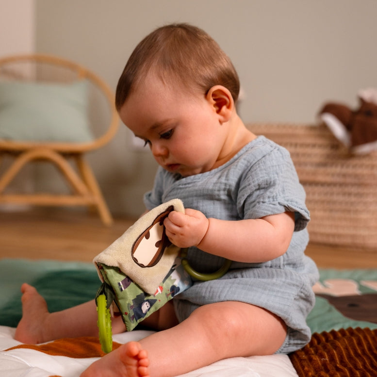A baby sitting on a play mat, holding a soft fabric sensory toy with a green handle. The baby is wearing a light blue muslin romper in a softly lit room with a wicker chair and basket in the background.