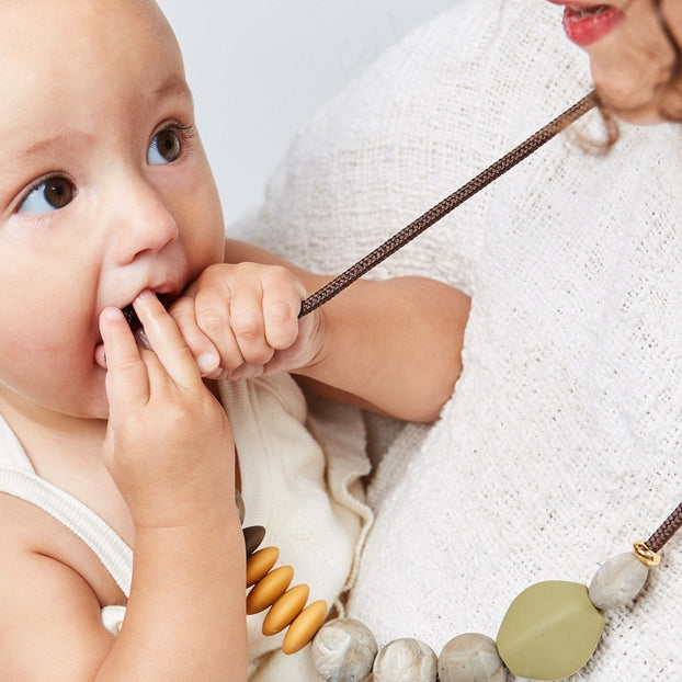 Woman holding a baby and wearing a necklace with large beads on a plain background.
