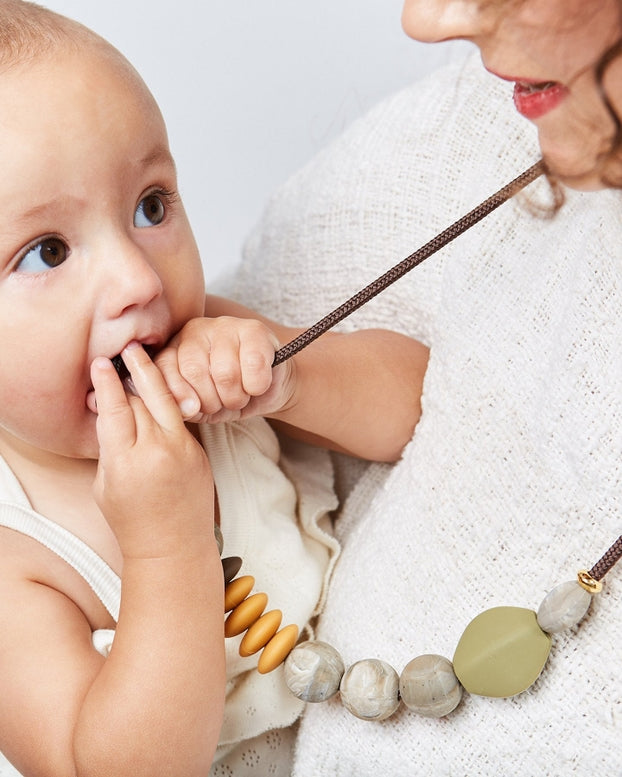 Woman holding a baby and wearing a necklace with large beads on a plain background.