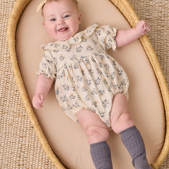 A smiling baby lying on a woven mat inside a natural wood bassinet, wearing a cream short-sleeve romper with a blue and yellow floral print, a matching floral headband, and gray ribbed booties.