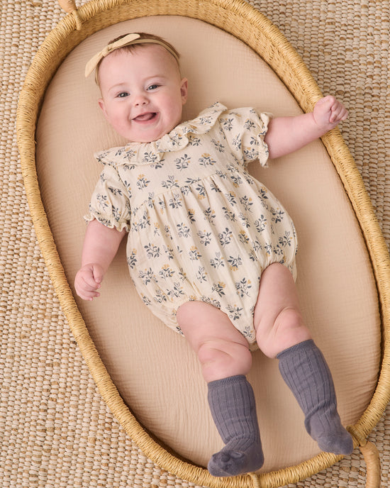 A smiling baby lying on a woven mat inside a natural wood bassinet, wearing a cream short-sleeve romper with a blue and yellow floral print, a matching floral headband, and gray ribbed booties.