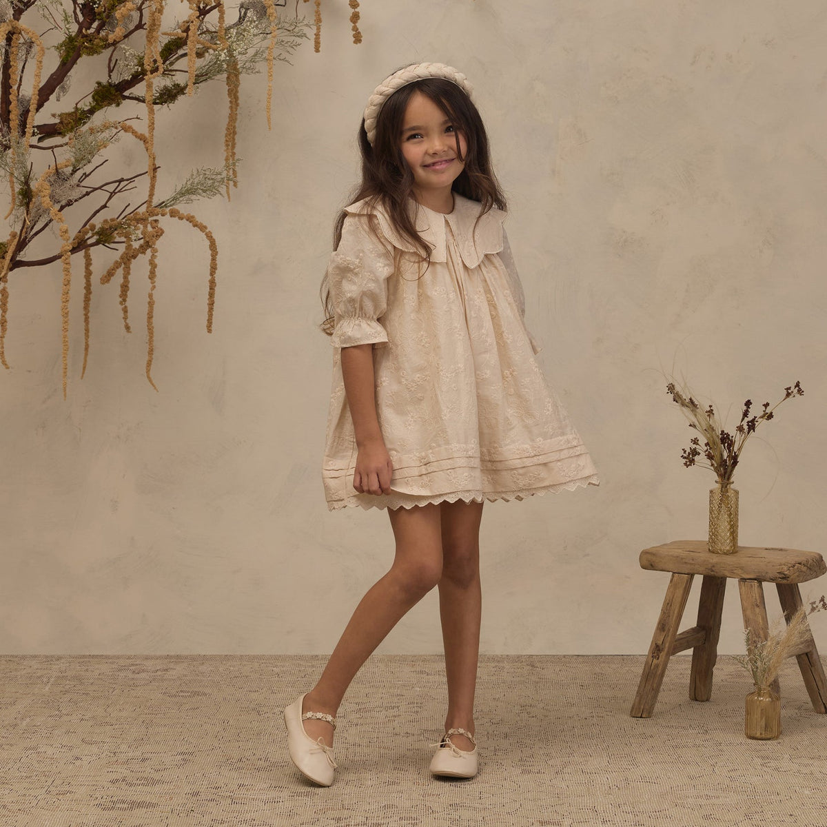 A young girl wearing a cream-colored lace dress with a matching headband and white shoes, standing in a styled studio setting with a textured beige wall, dried floral decorations, and a small wooden stool.