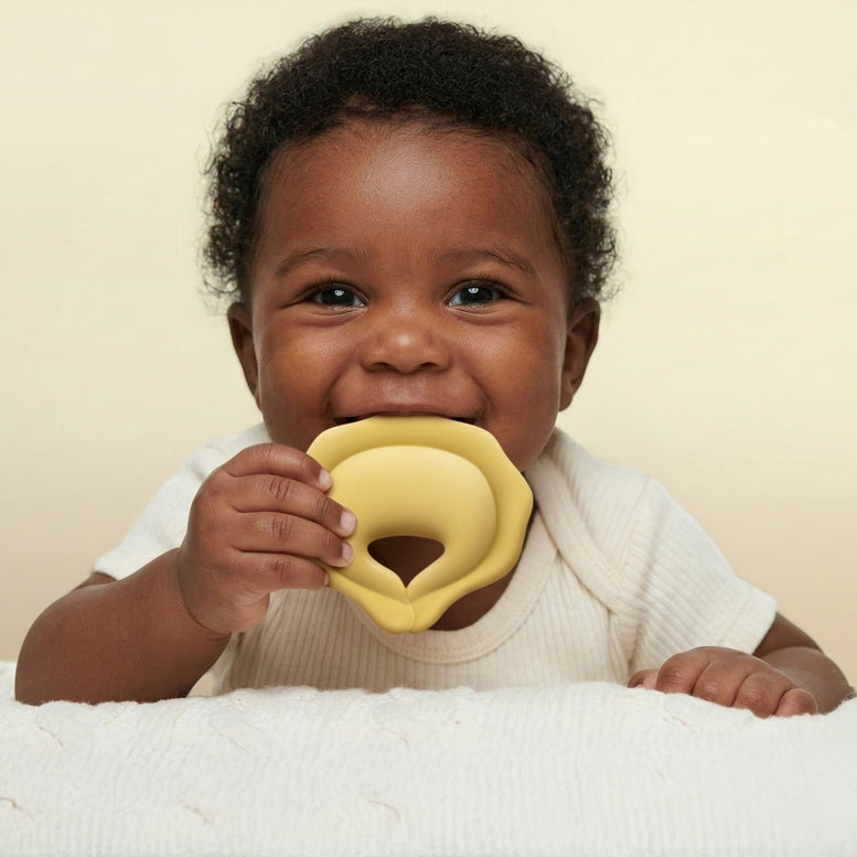 A smiling baby with dark curly hair wearing a white ribbed onesie, holding a yellow silicone teether in their mouth against a warm cream background.