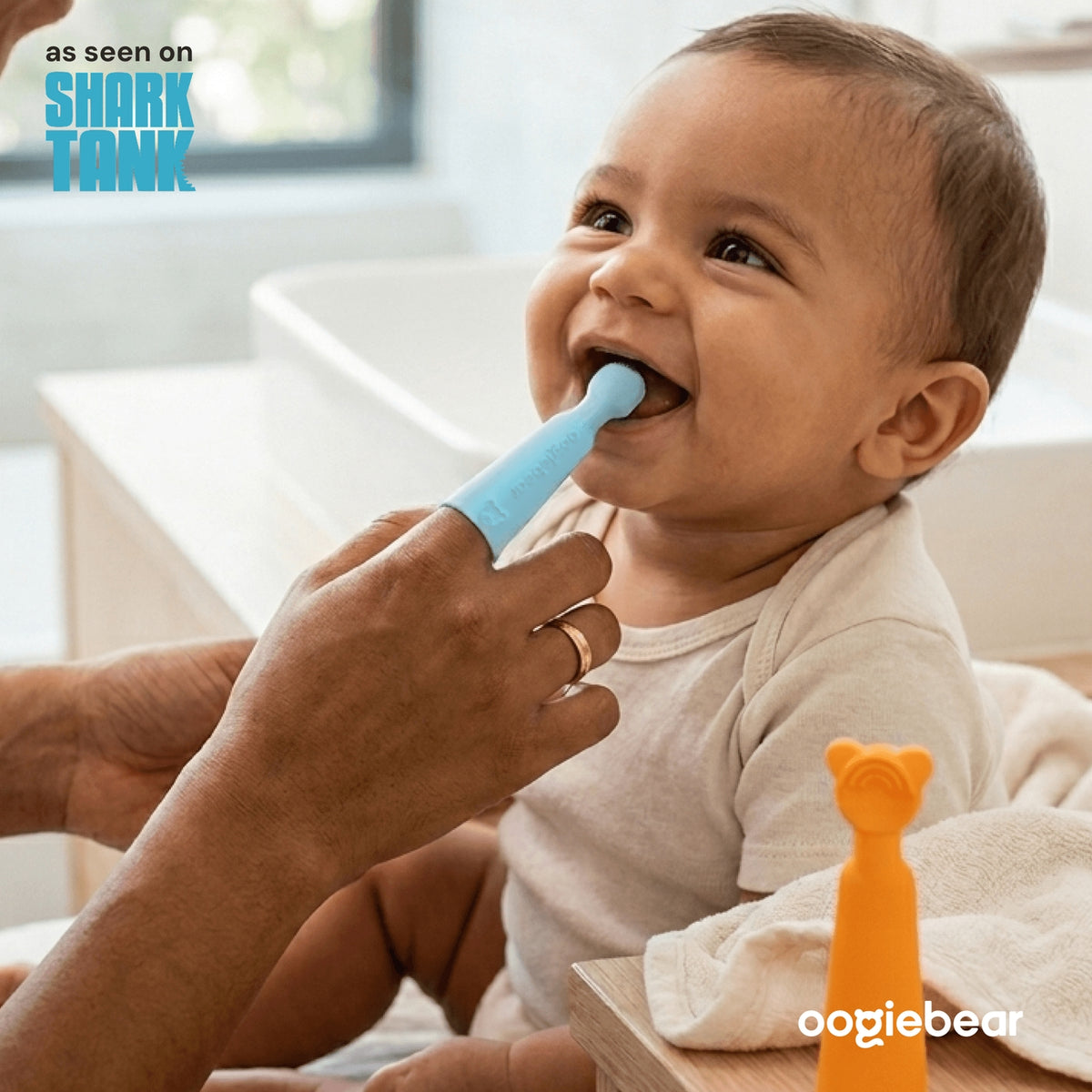 Baby being assisted with a blue toothbrush by an adult, with 'Shark Tank' branding and 'oogiebear' product visible.