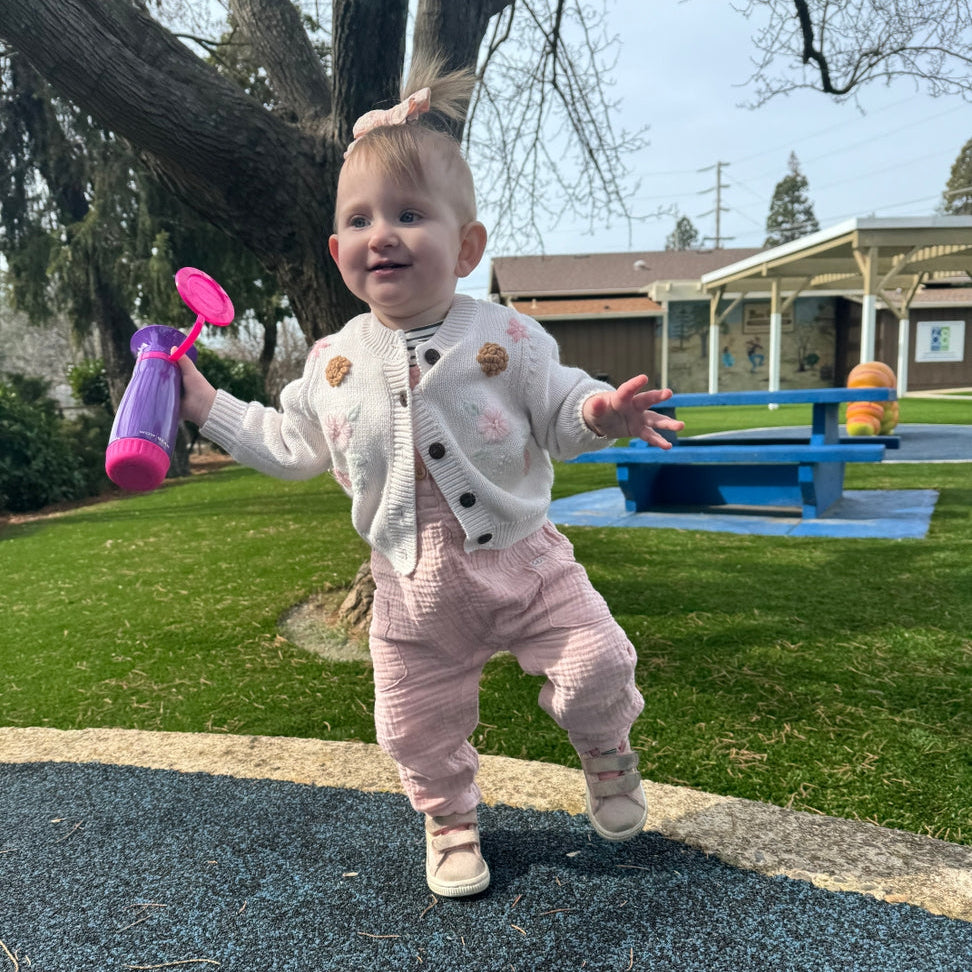 A toddler walking outdoors on a playground, wearing a cream cardigan with pink polka dots and light pink pants, holding a pink and purple toy.