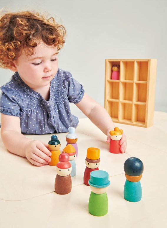A toddler with curly hair sits at a light wood table playing with colorful wooden peg dolls, with a small wooden storage box in the background.