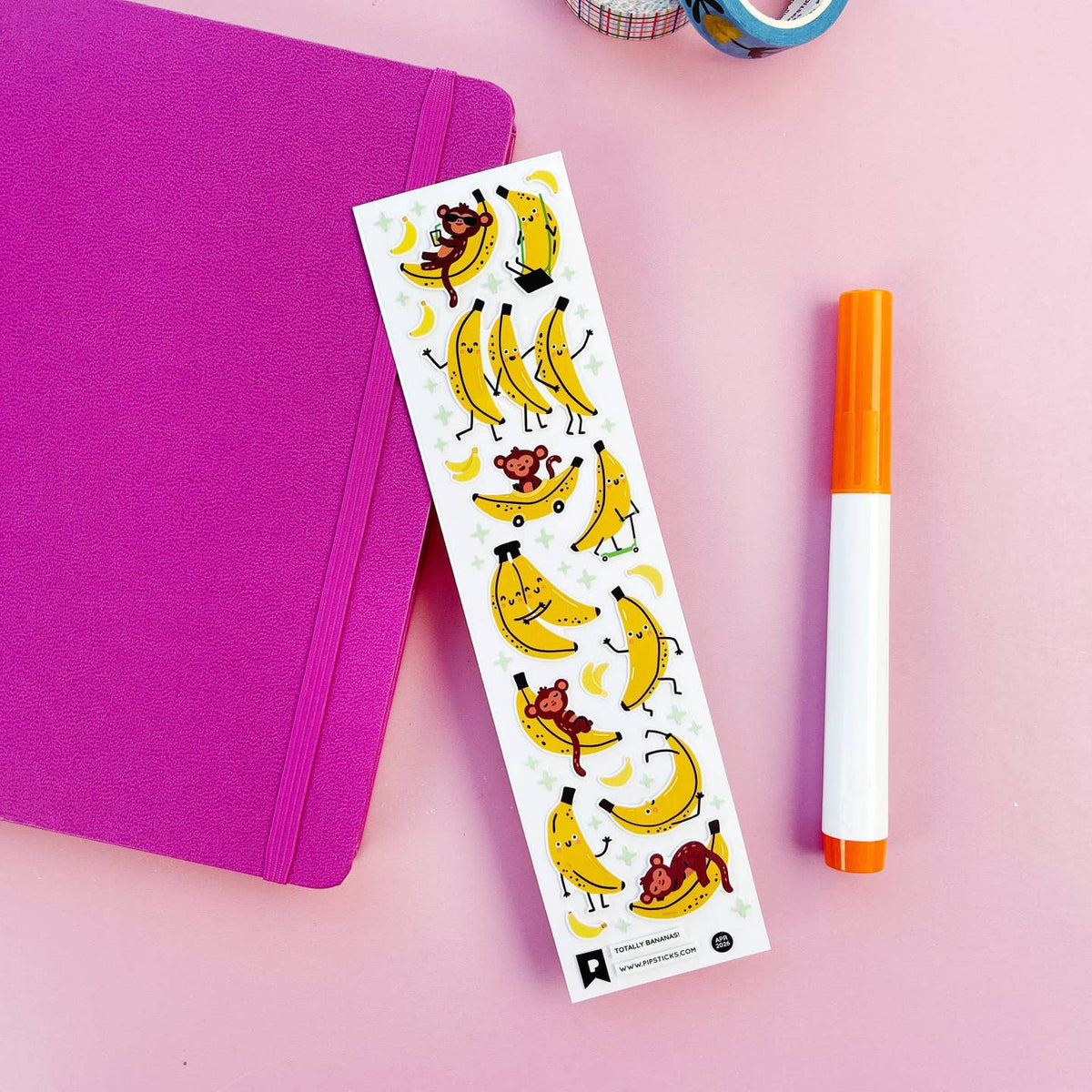 Sheet of banana-themed stickers with cartoon monkeys on a pink surface with a purple notebook and orange marker.