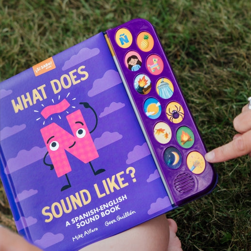 A person holding "What Does Ñ Sound Like?: A Spanish-English Sound Book" outdoors on grass, showing the purple cover with an animated letter Ñ character, clouds, and a vertical strip of interactive sound buttons.