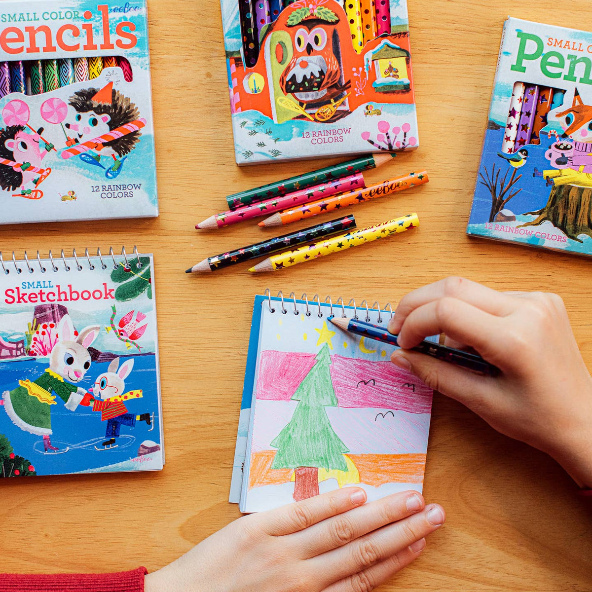 A child's hands are shown drawing in a small sketchbook with a blue pencil on a wooden table, surrounded by colorful children's art books and loose colored pencils.