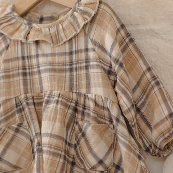 A close-up of a children's plaid dress in beige, brown, and gray tones, hanging on a wooden hanger against a textured beige fabric background.